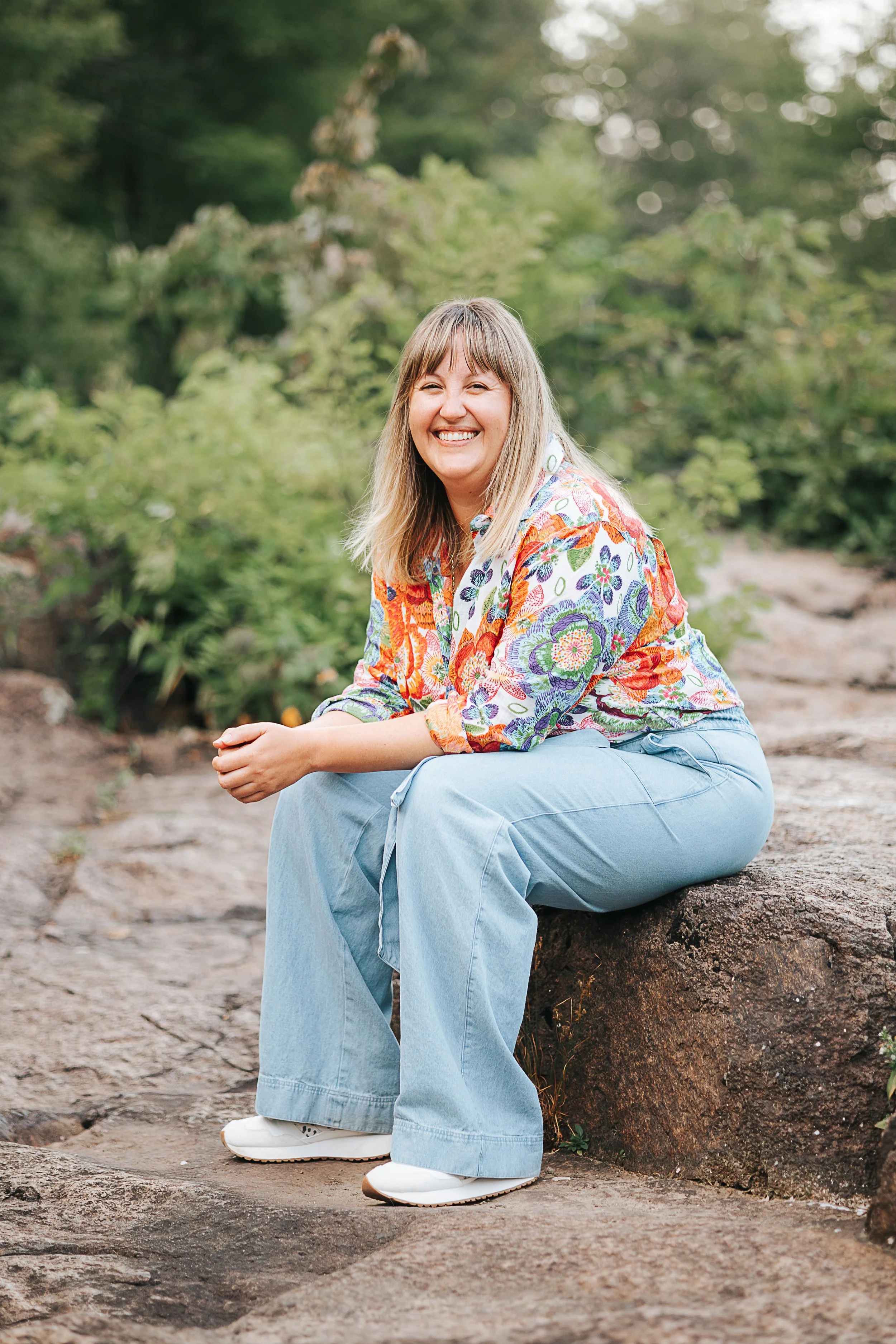 Une femme souriante, assise sur une roche dans un environnement naturel verdoyant.