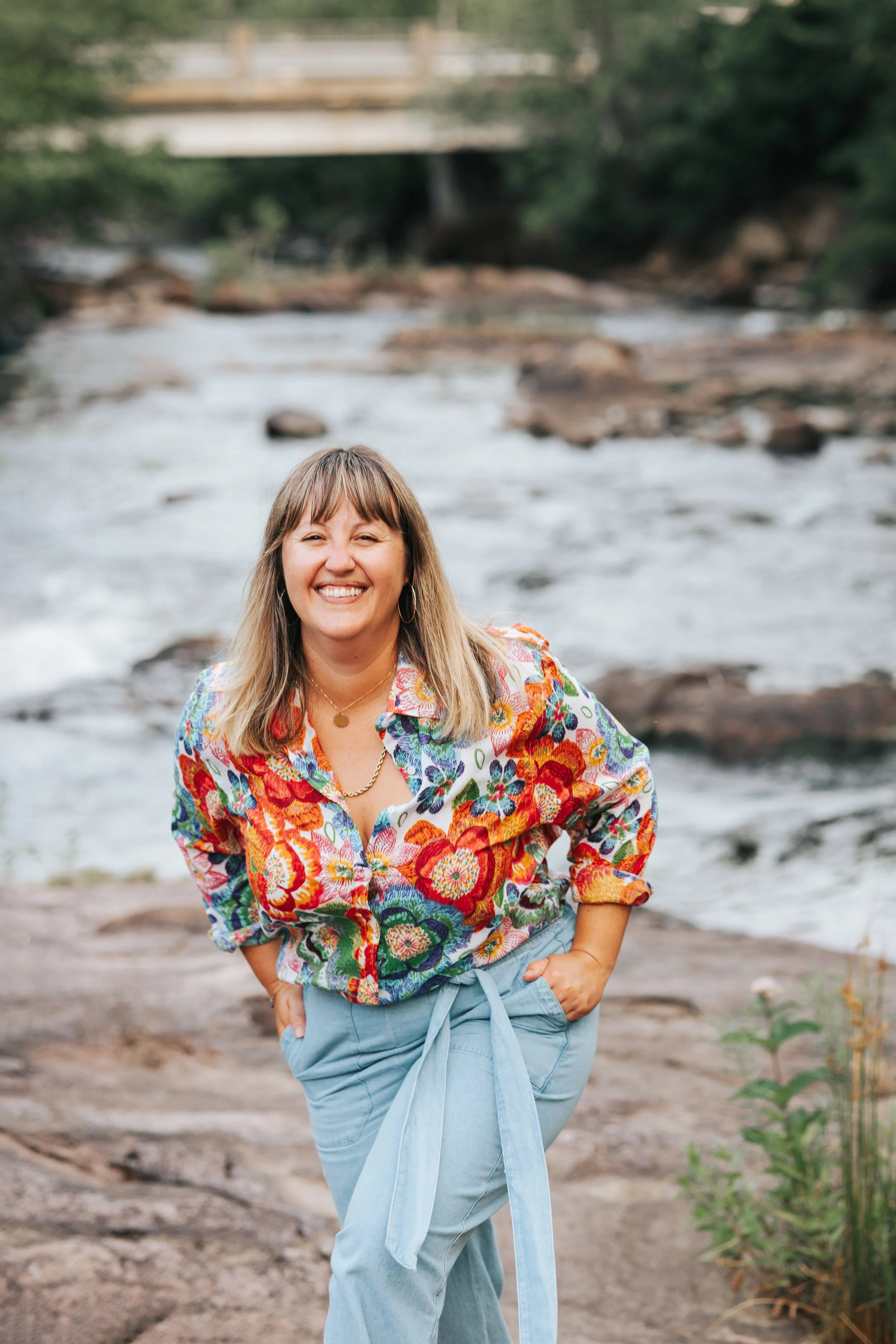Une femme souriante portant une chemise colorée à fleurs et un pantalon bleu, debout près d'une rivière avec un pont en arrière-plan.