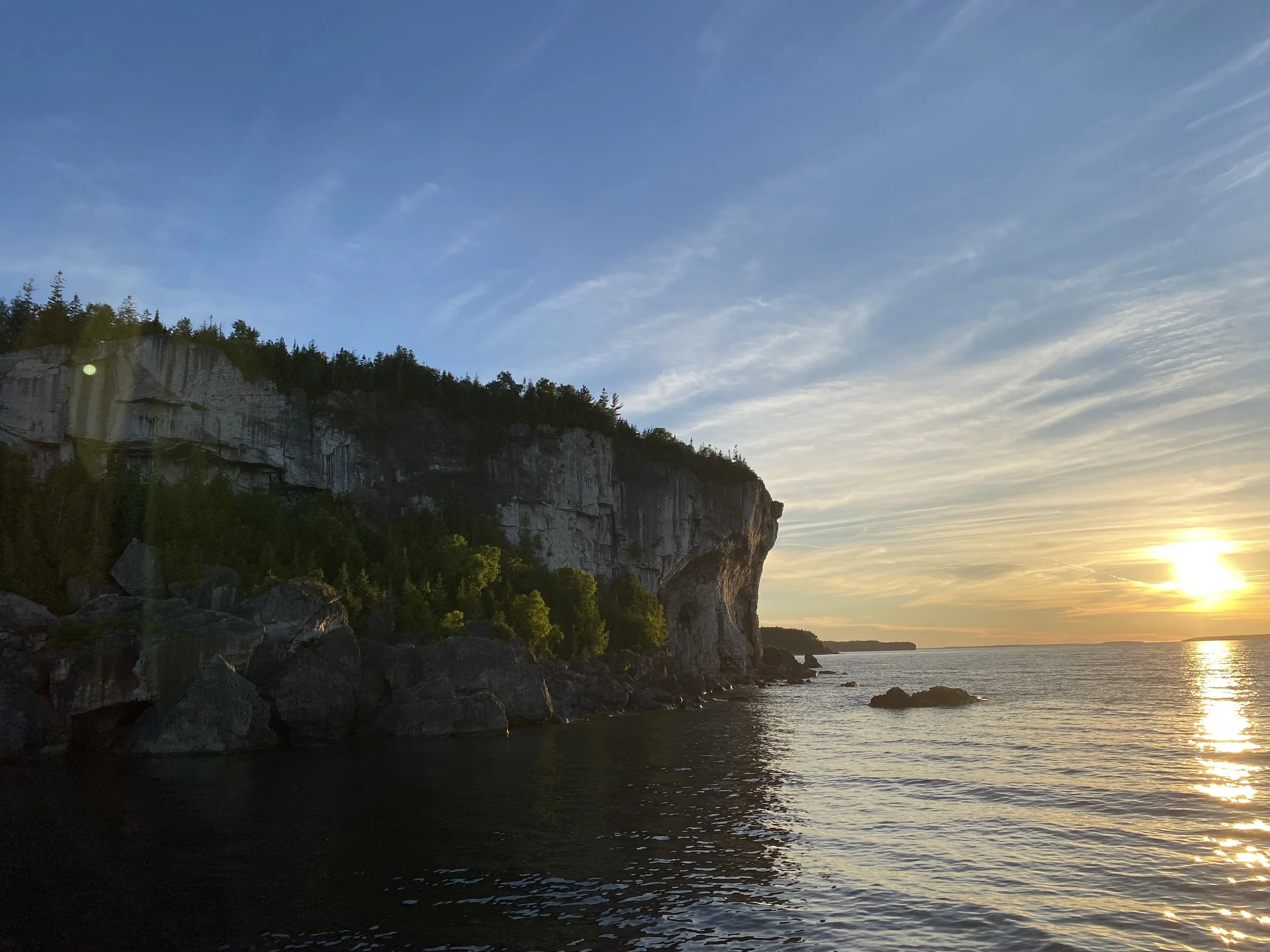 Coucher de soleil sur une mer calme avec des falaises escarpées et des rochers, ciel dégagé avec quelques nuages.