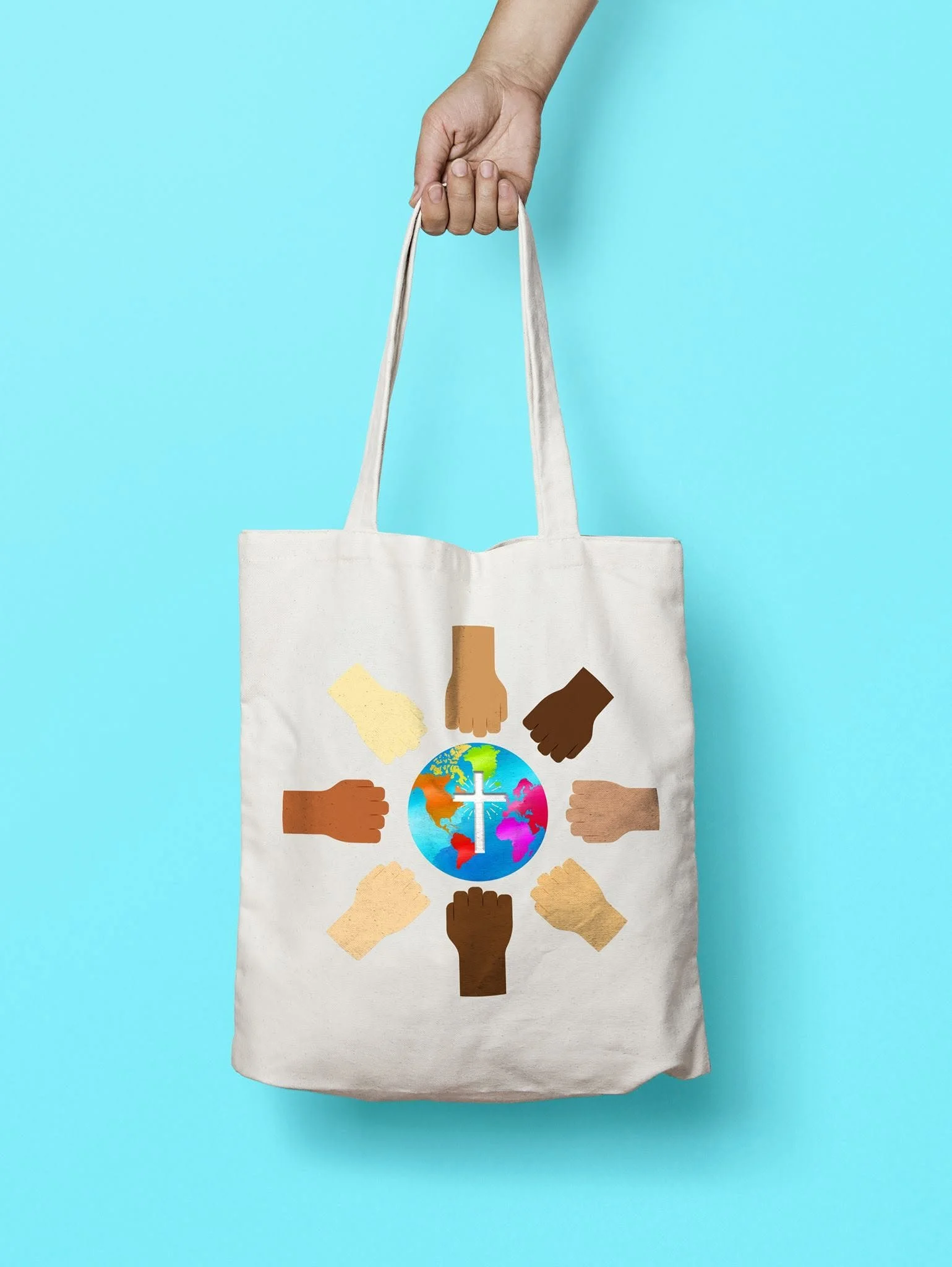 A white tote bag with illustrations of diverse hands surrounding a globe with a cross on it, set against a light blue background. A hand is holding the bag's handles.