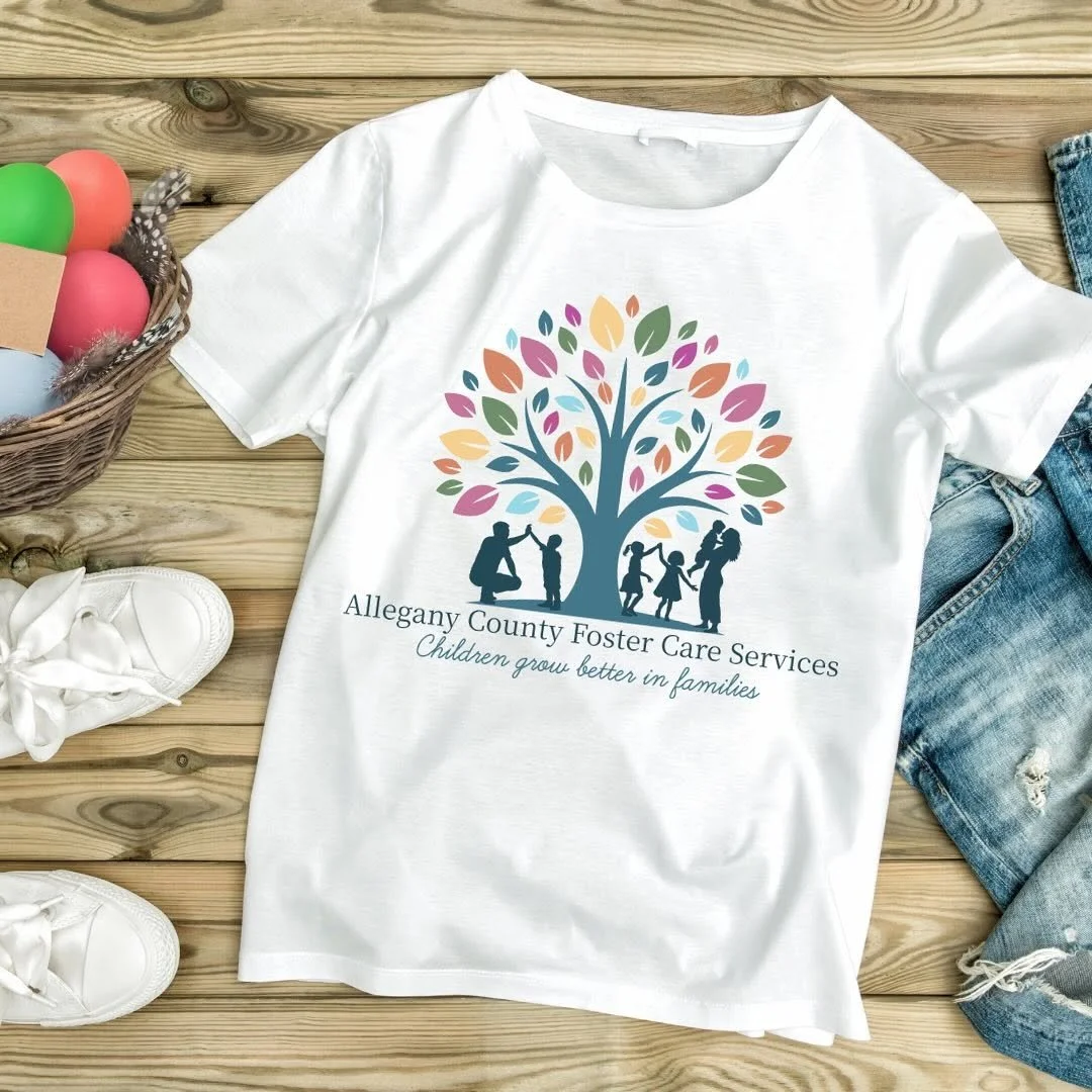White T-shirt with a colorful tree and children playing design, promoting Allegany County Foster Care Services, placed on a wooden surface with sneakers, jeans, and Easter eggs nearby.