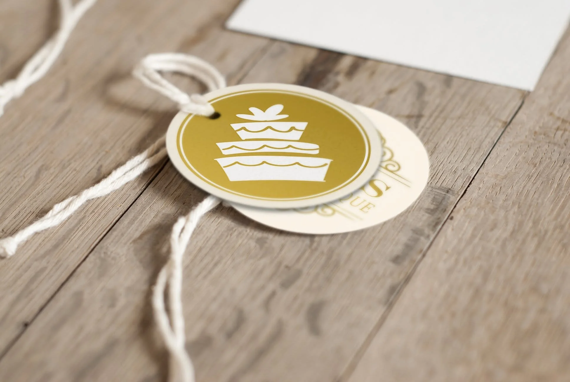 A round, gold-colored tag with a white birthday cake icon and a bow on top, attached to a piece of white string, lying on a wooden surface with a white document in the background.