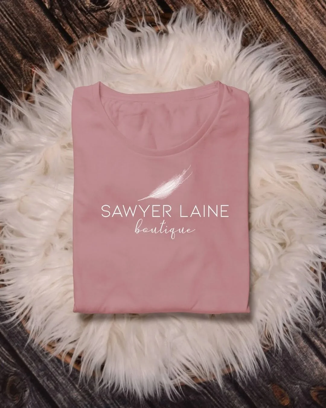 Pink T-shirt with 'Sawyer Laine boutique' written on it, placed on a white fluffy rug on a wooden surface.