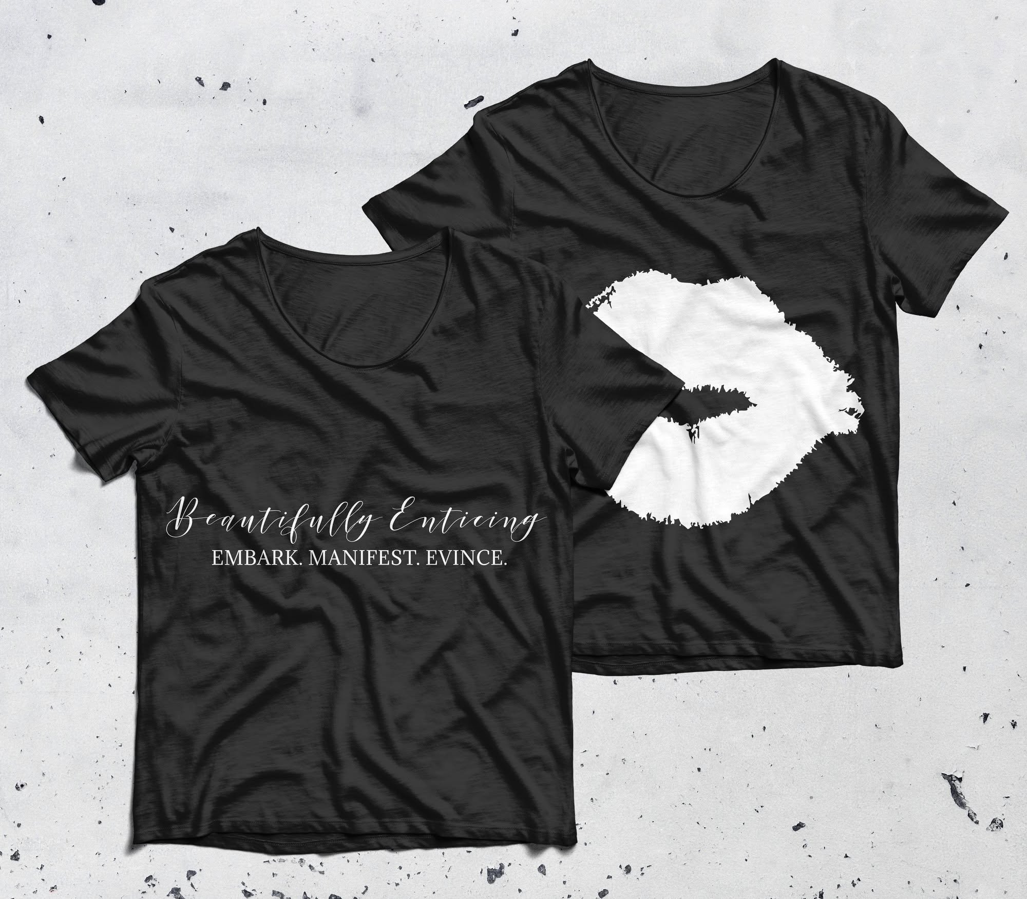Black T-shirts with a white outline of a person's face and text that reads 'Beautifully Enticing, EMBARK. MANIFEST. EVINCE.' on a gray surface with small black paint splatters.