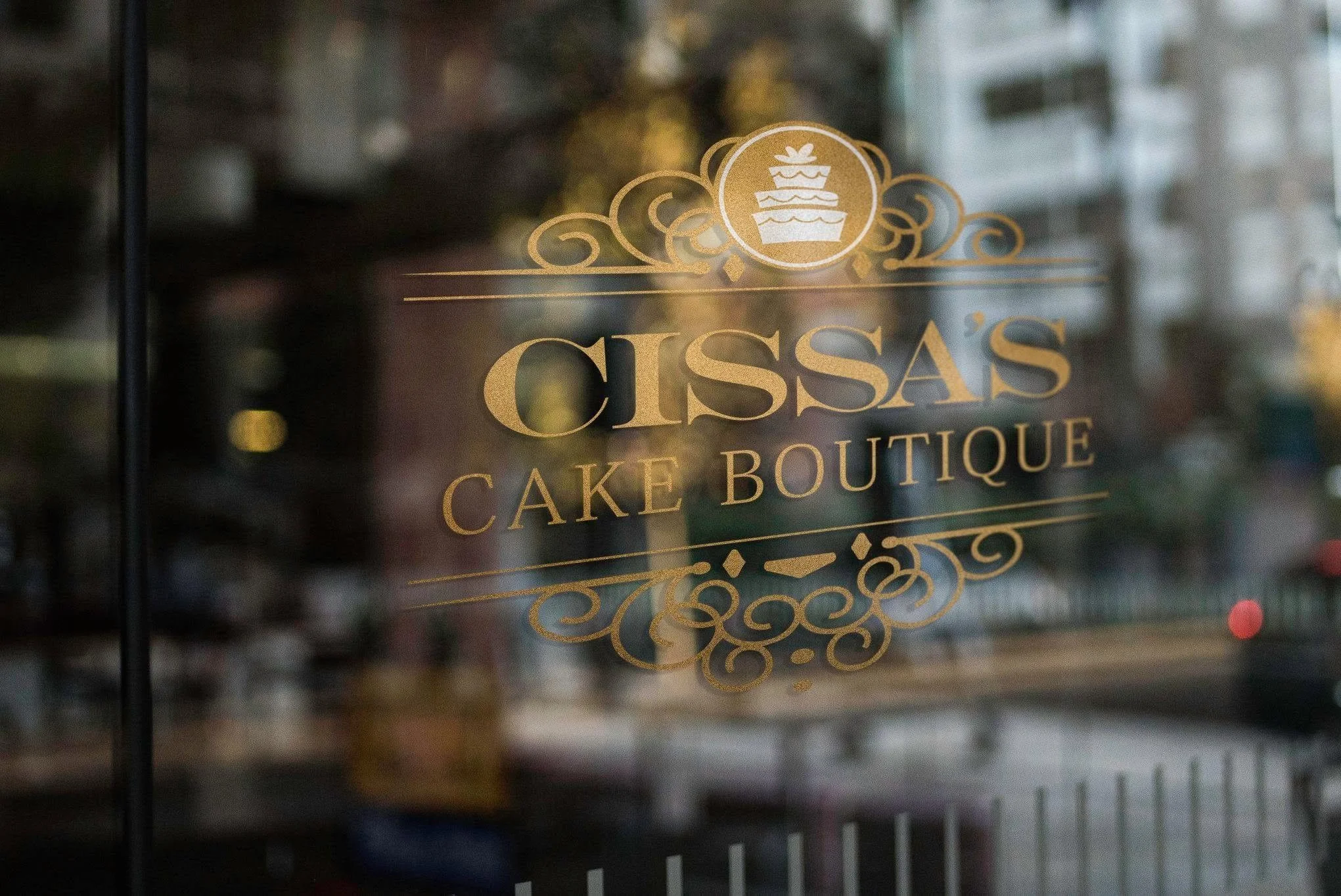 Glass window with gold lettering and ornate design reading 'Cissa's Cake Boutique' with a cupcake logo above.