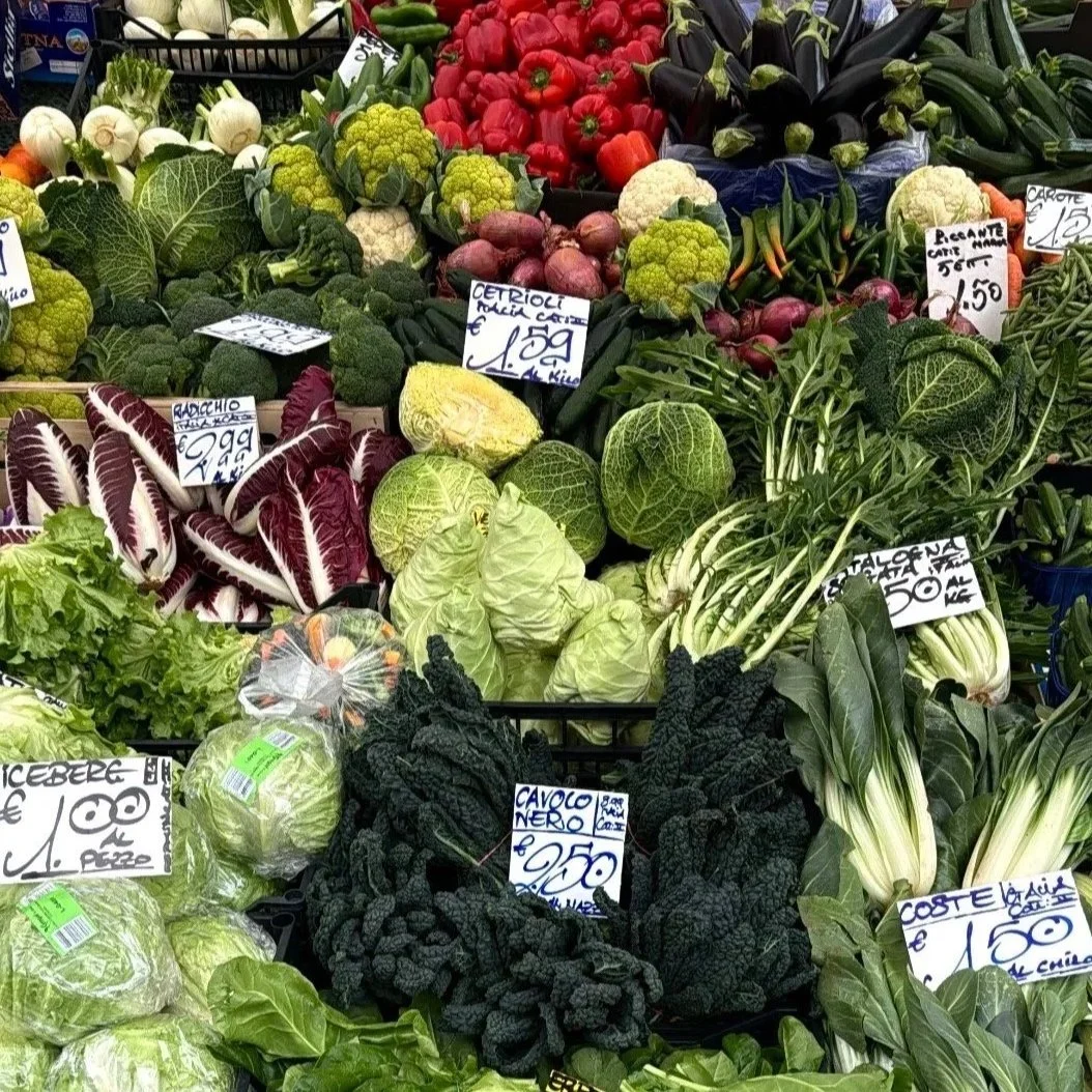 Display of fresh vegetables at a market, including calabrese, lettuce, radicchio, broccoli, cauliflower, cucumbers, and tomatoes, with handwritten price tags in Italian.