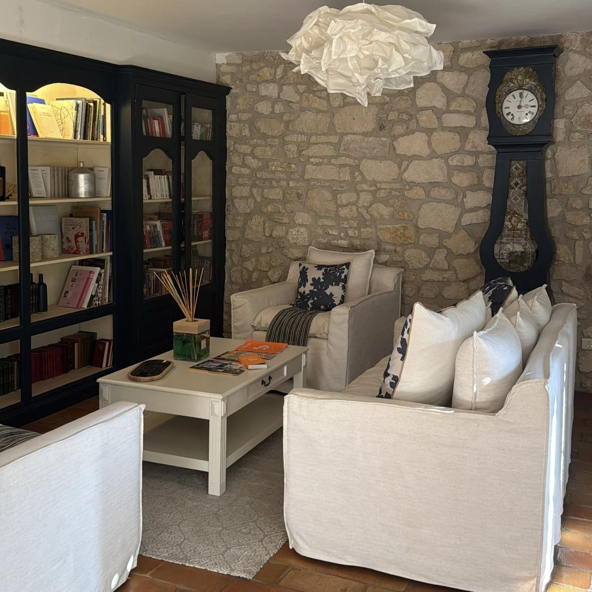 Living room with stone wall, white sofa and armchair, black clock, and bookshelf, with a paper lamp overhead.