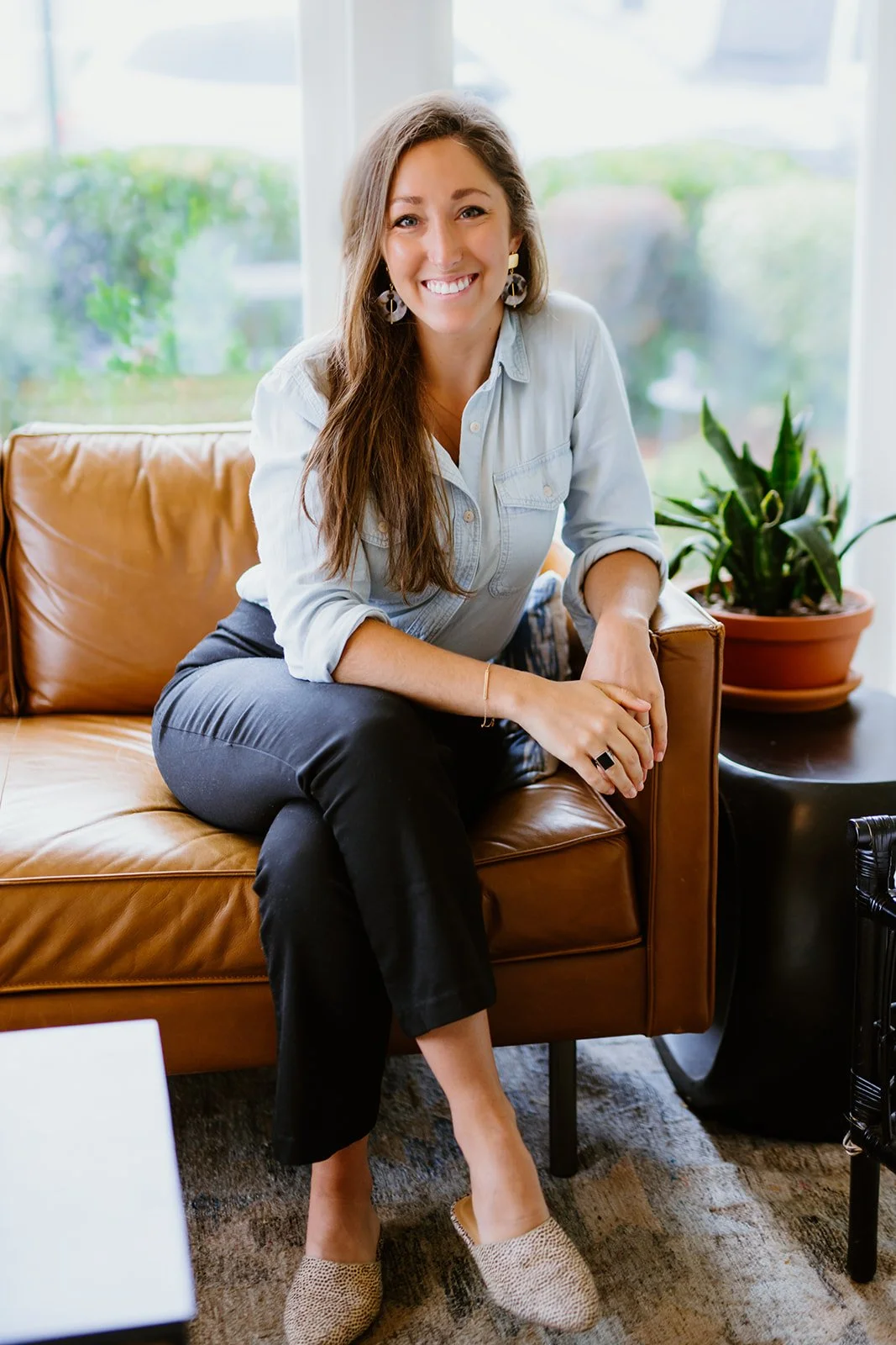 A woman with long brown hair sitting on a tan leather sofa in a bright room with a large window and a potted plant in the background. She is smiling and looking at the camera, wearing a light blue shirt, black pants, and beige shoes.