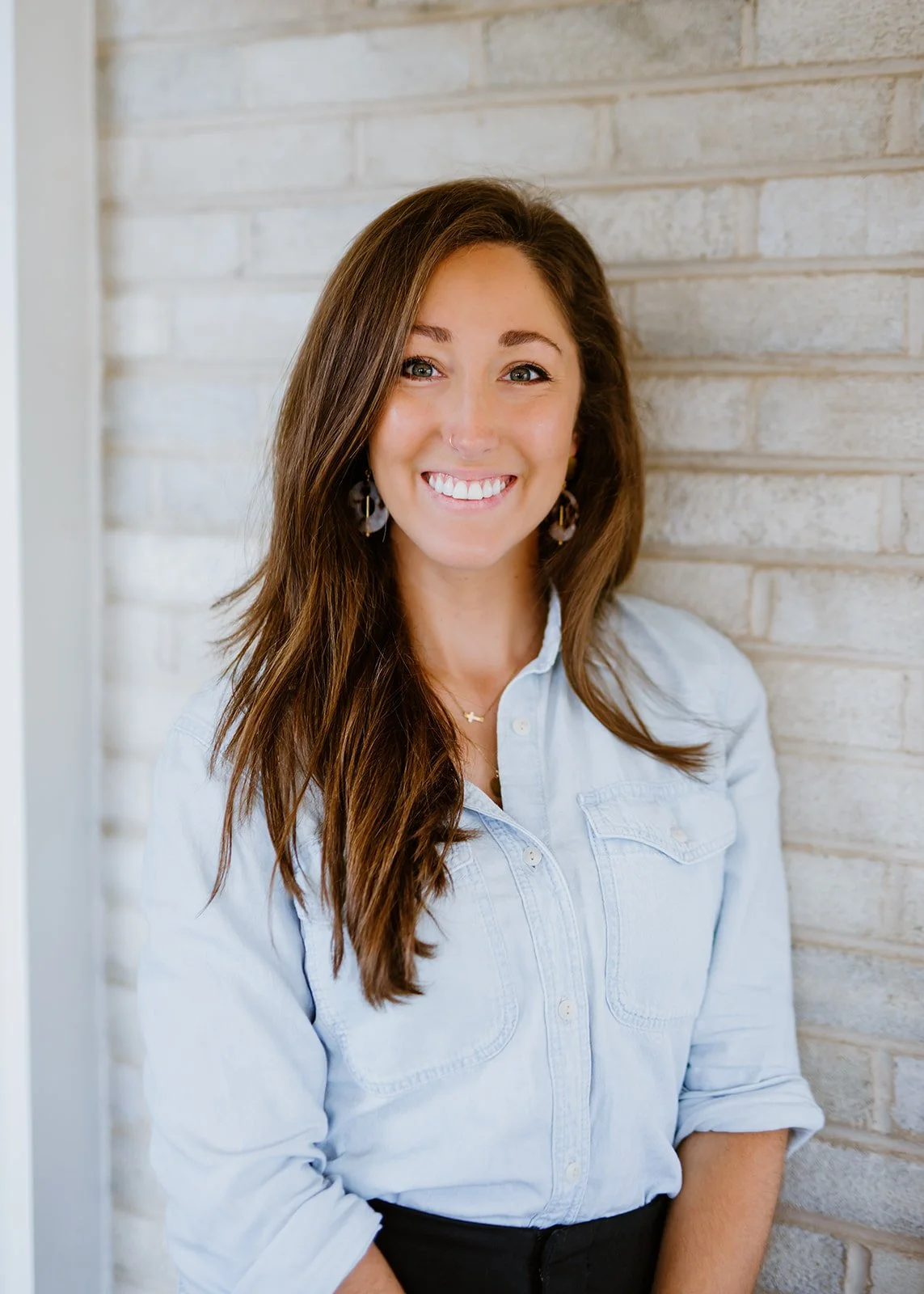 A woman with long brown hair, smiling, wearing a white button-up shirt, earrings, and a necklace, standing against a light-colored brick wall.