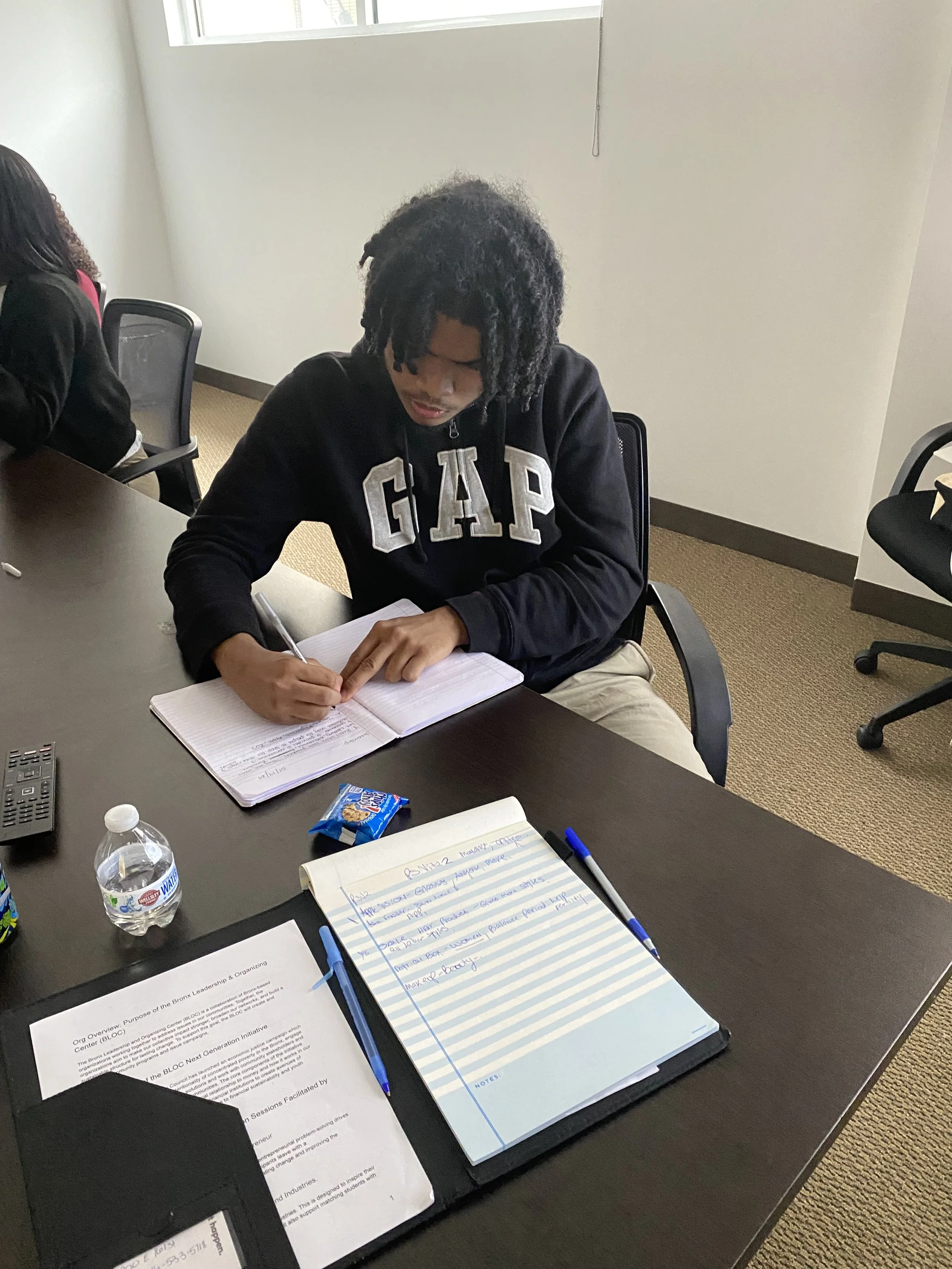 Person with curly hair wearing a black GAP hoodie sitting at a desk, writing in a notebook, with documents, a water bottle, a snack, and several pens on the desk.