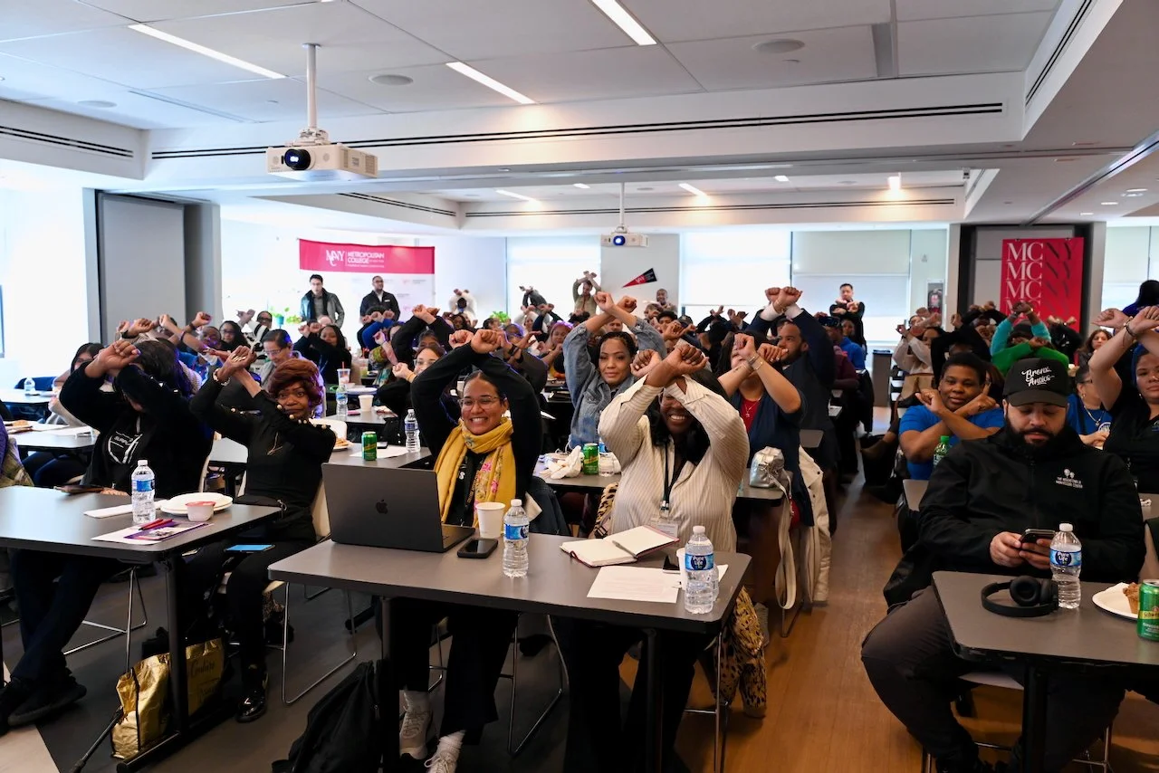 A large group of smiling people sitting at desks in a conference room, crossing their arms above their heads in an "X" for the Bronx, with some using laptops and drinking water bottles, participating in The Bronx Does Not Back Down forum.
