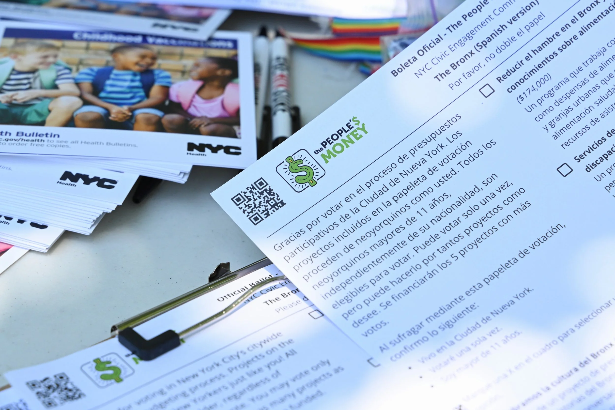 English and Spanish official NYC ballot paper with a QR code, printed text, and a checkbox, surrounded by informational pamphlets and pencils.