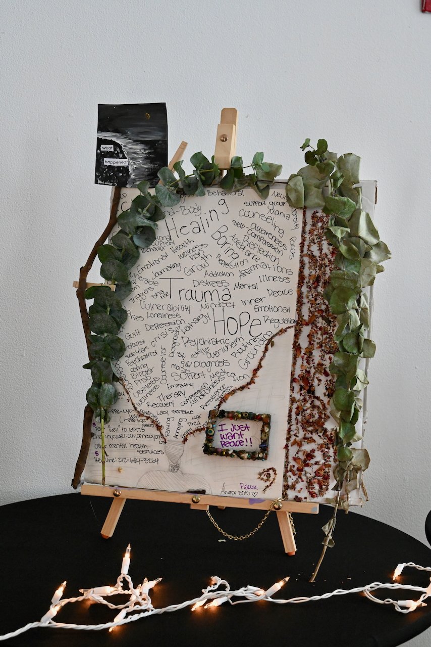 A poster on an easel with the word 'trauma' crossed out and replaced with 'hope', surrounded by handwritten notes about mental health and healing. Decorated with green foliage and string lights.