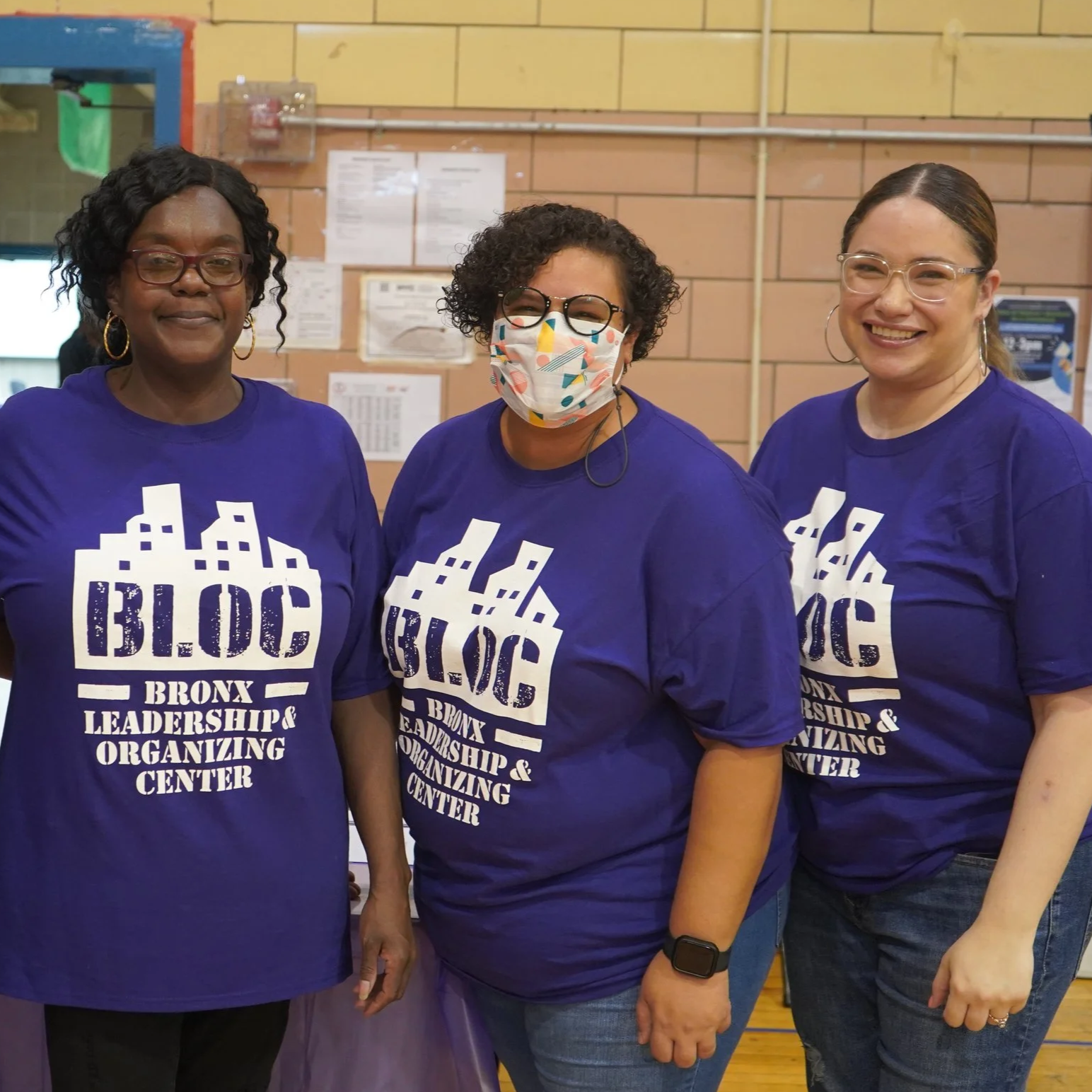 Three women wearing matching purple T-shirts with a cityscape and text that reads "BLOC Bronx Leadership & Organizing Center," standing together indoors.