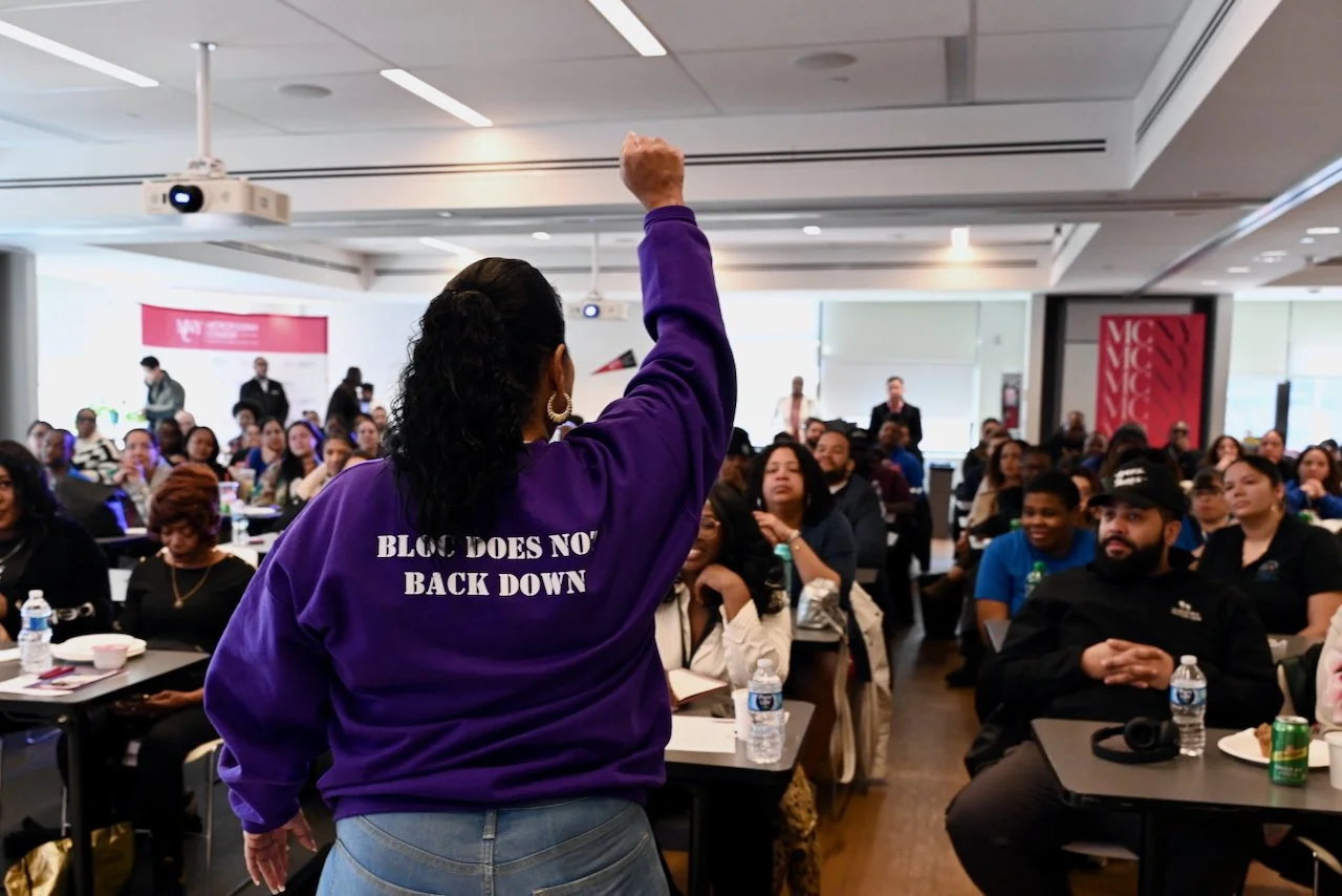A woman with a purple sweatshirt that says 'BLOC DOES NOT BACK DOWN' standing in front of an audience in a conference room, raising her fist in the air as she addresses the crowd.