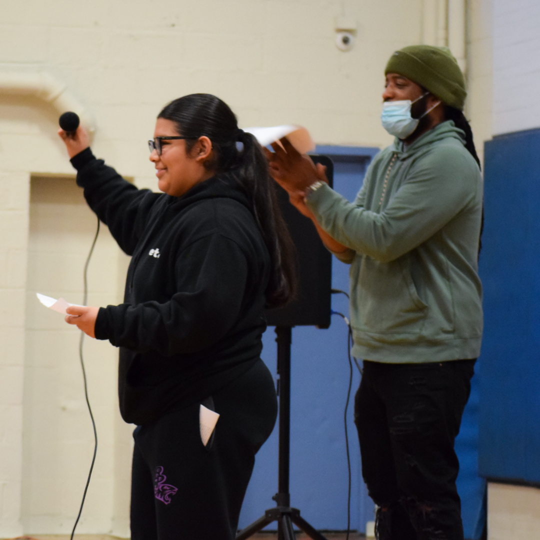 Two people stand on a stage, one speaking into a microphone and the other clapping, inside a gym with a blue padded wall in the background.