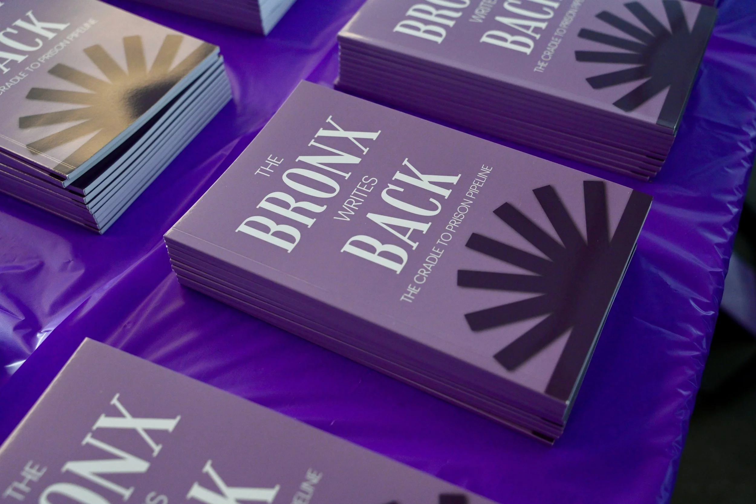 Stacks of purple booklets titled 'The Bronx Writes Back: The Cradle to Prison Pipeline' placed on a purple tablecloth.