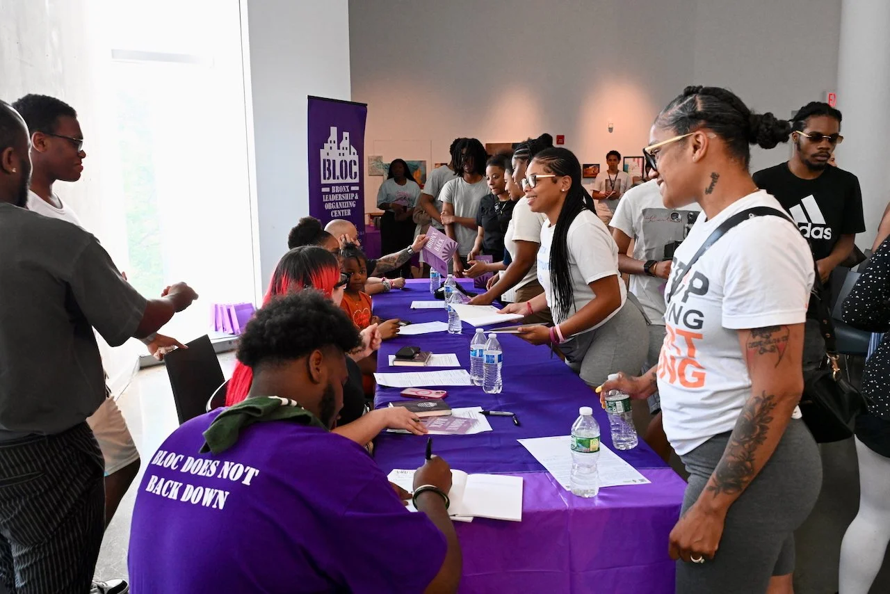 People asking youth authors to sign their copies of The Bronx Writes Back at a book launch, with some sitting and others standing in line, at a table with purple tablecloths, bottles of water, and papers, in a well-lit room with artwork on the walls.