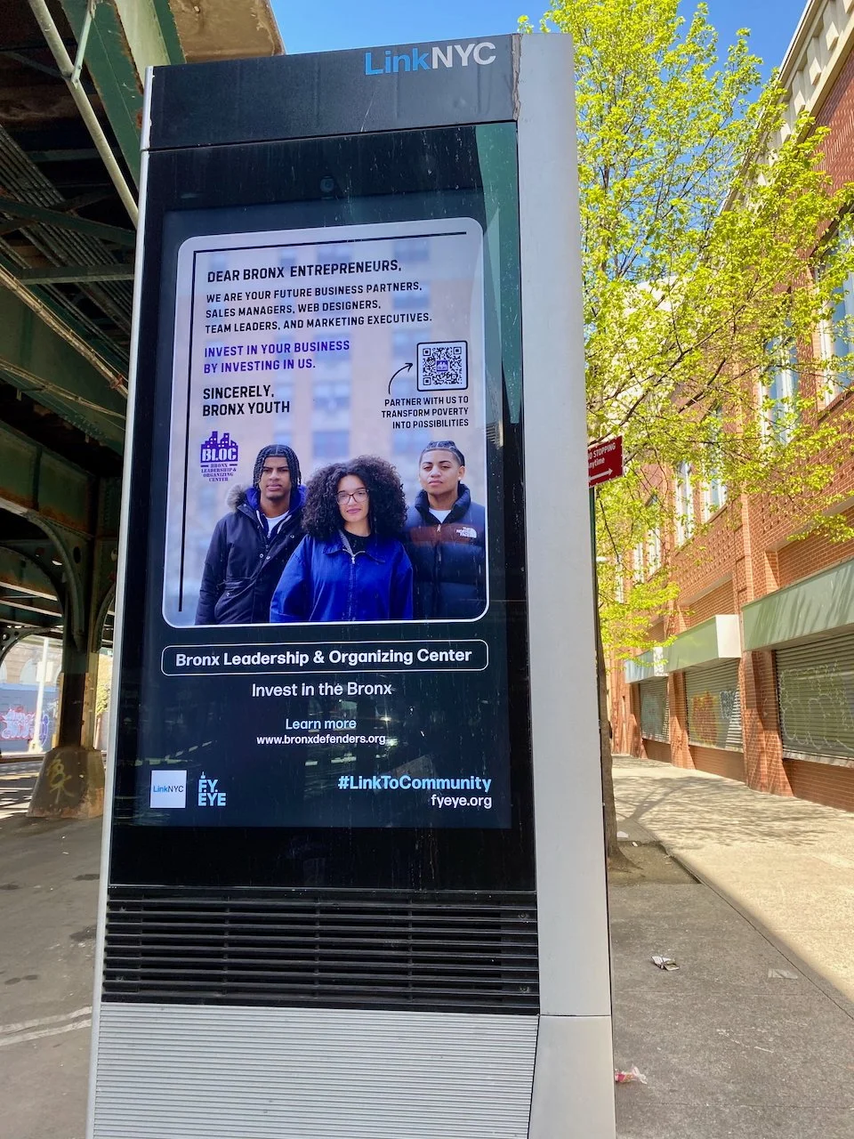 A digital billboard on a city sidewalk with an advertisement from LinkNYC for Bronx youth entrepreneurs. The ad features a photo of three young people in blue jackets and text encouraging business investment and community involvement. The billboard is under an elevated train track with a building and trees nearby.