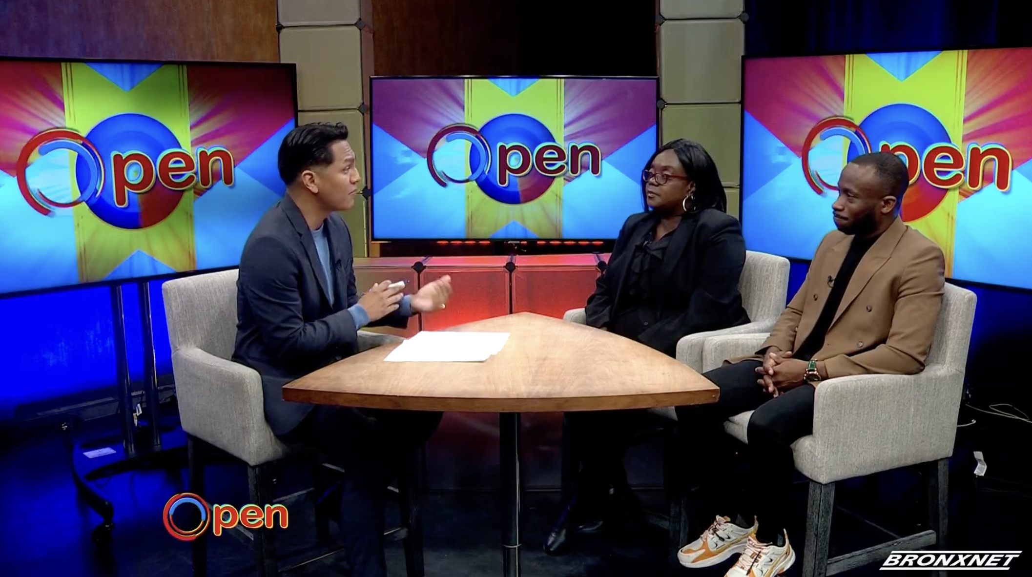 Three people in a television studio sitting at a round wooden table during a talk show. Behind them are three large screens displaying the word 'Open' with colorful background graphics. The person on the left is a man in a dark blazer and shirt, speaking and gesturing with his hands. The woman in the middle is wearing glasses, a black blazer, and has shoulder-length hair. The man on the right is dressed in a tan blazer, black shirt, and colorful sneakers. The studio has a professional setting with a mix of warm and cool lighting.