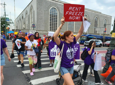 A group of people participating in a protest march on a city street, holding signs advocating to freeze rent now.