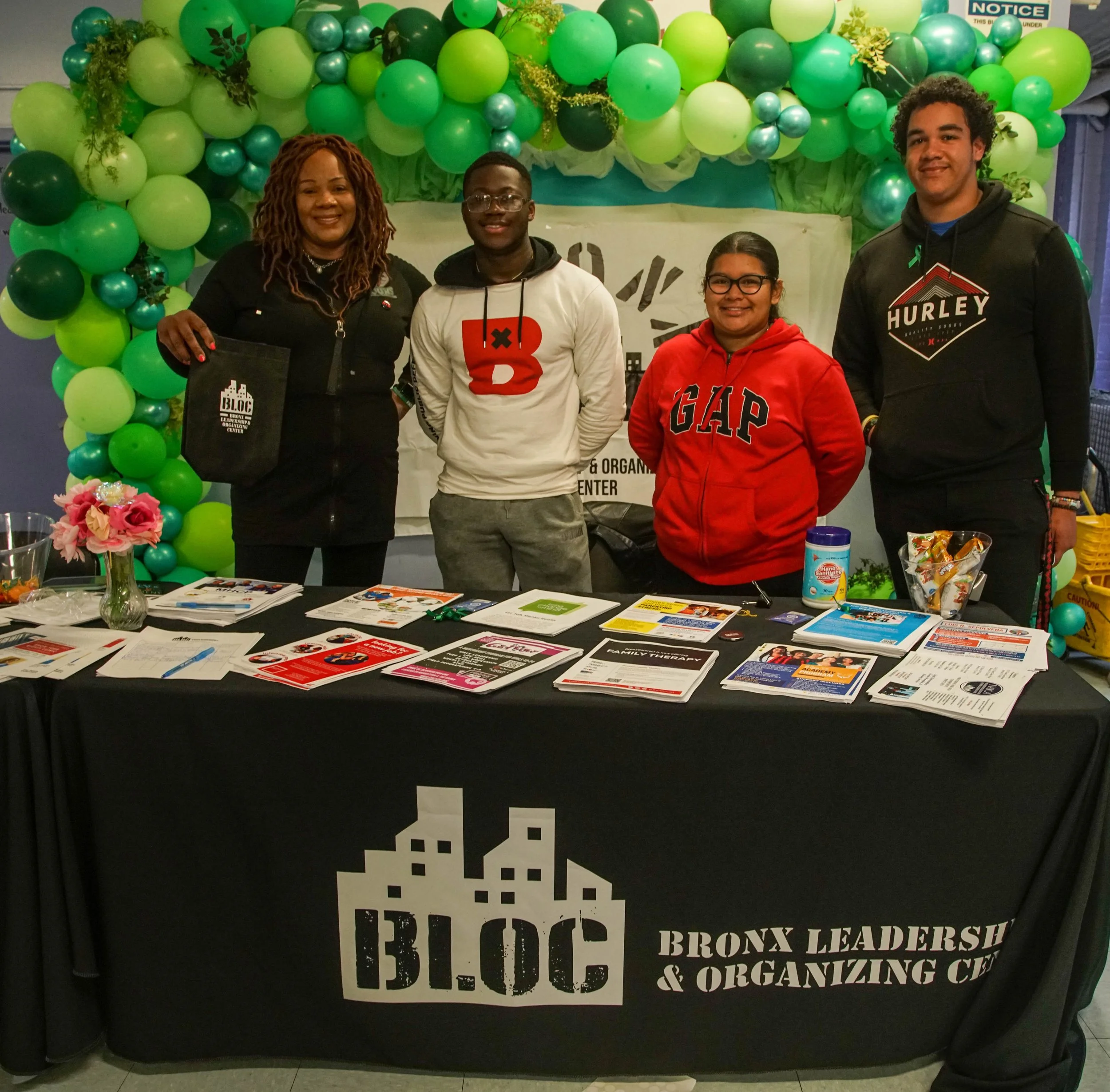 Group of four people standing behind a table covered with brochures, flyers, and pamphlets at a Bronx Leadership & Organizing Center event, with an arch of green balloons in the background.