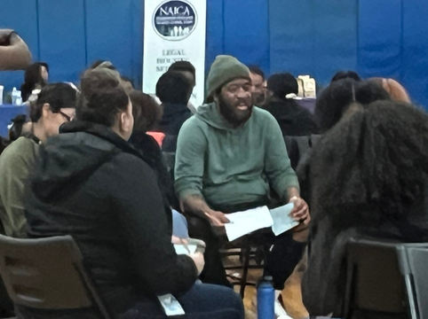 A group of people sitting in chairs in a circle, engaged in a discussion, with a man holding papers smiling in the center.