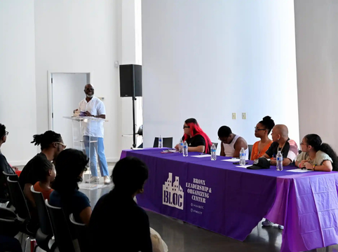 A woman is speaking at a podium in front of an audience at The Bronx Writes Back book launch. The table has a purple tablecloth with the words 'Bronx Leadership & Organizing Center' and 'BLOC' written on it, and water bottles are placed on the table.