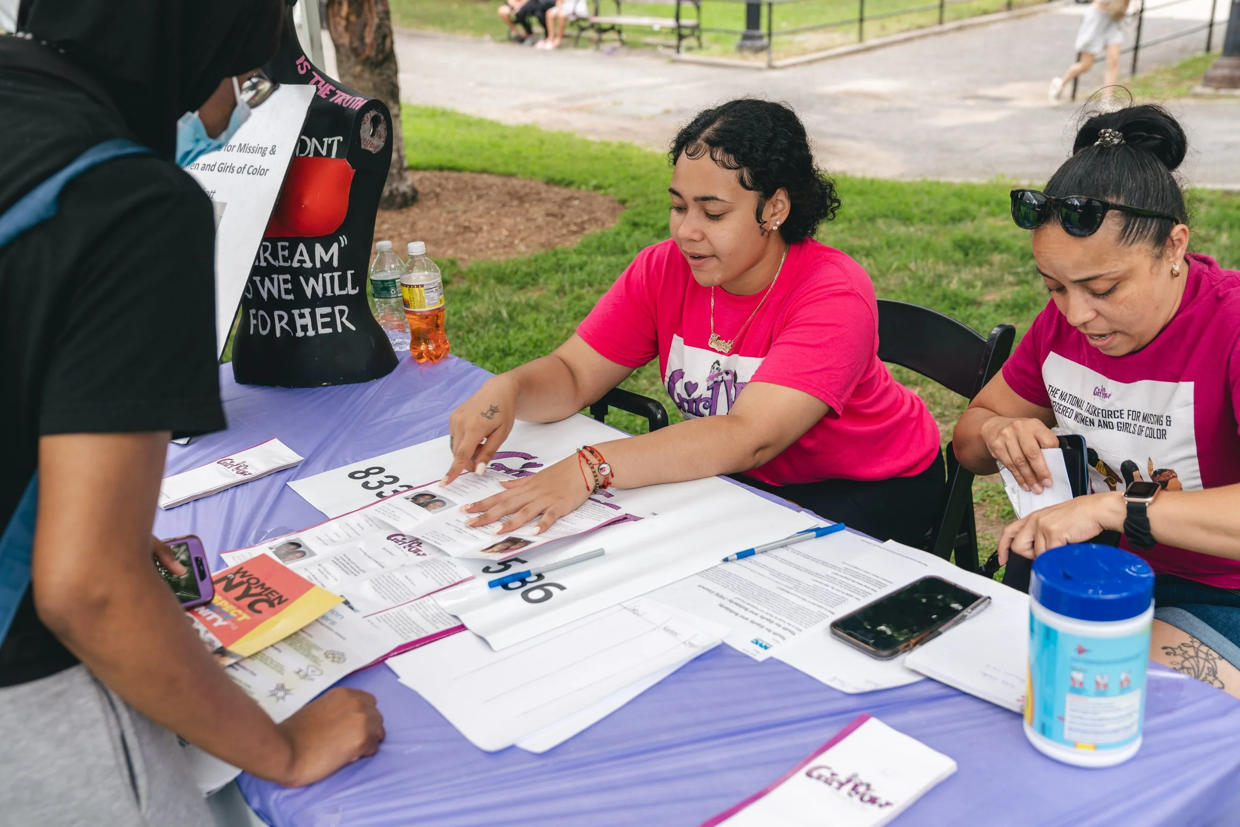 Two women sitting at a table outdoors, engaging with a person standing in front of them, at a community fair. The women are wearing pink shirts, and the table has flyers, brochures, and informational materials. One woman is pointing at a brochure, and there are water bottles and a container on the table.