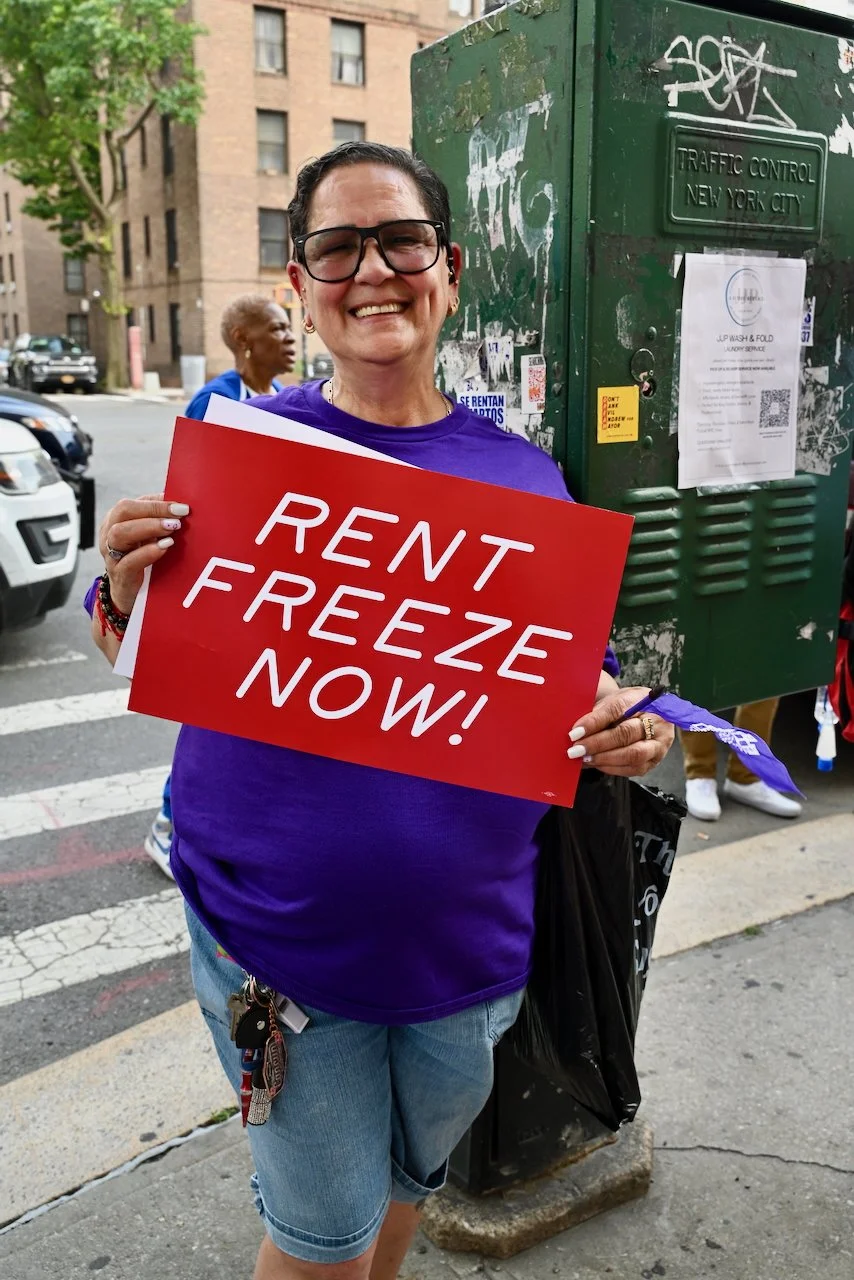 A woman smiling and holding a red sign that reads 'RENT FREEZE NOW!' on a city street with cars, pedestrians, and a green traffic control box in the background.