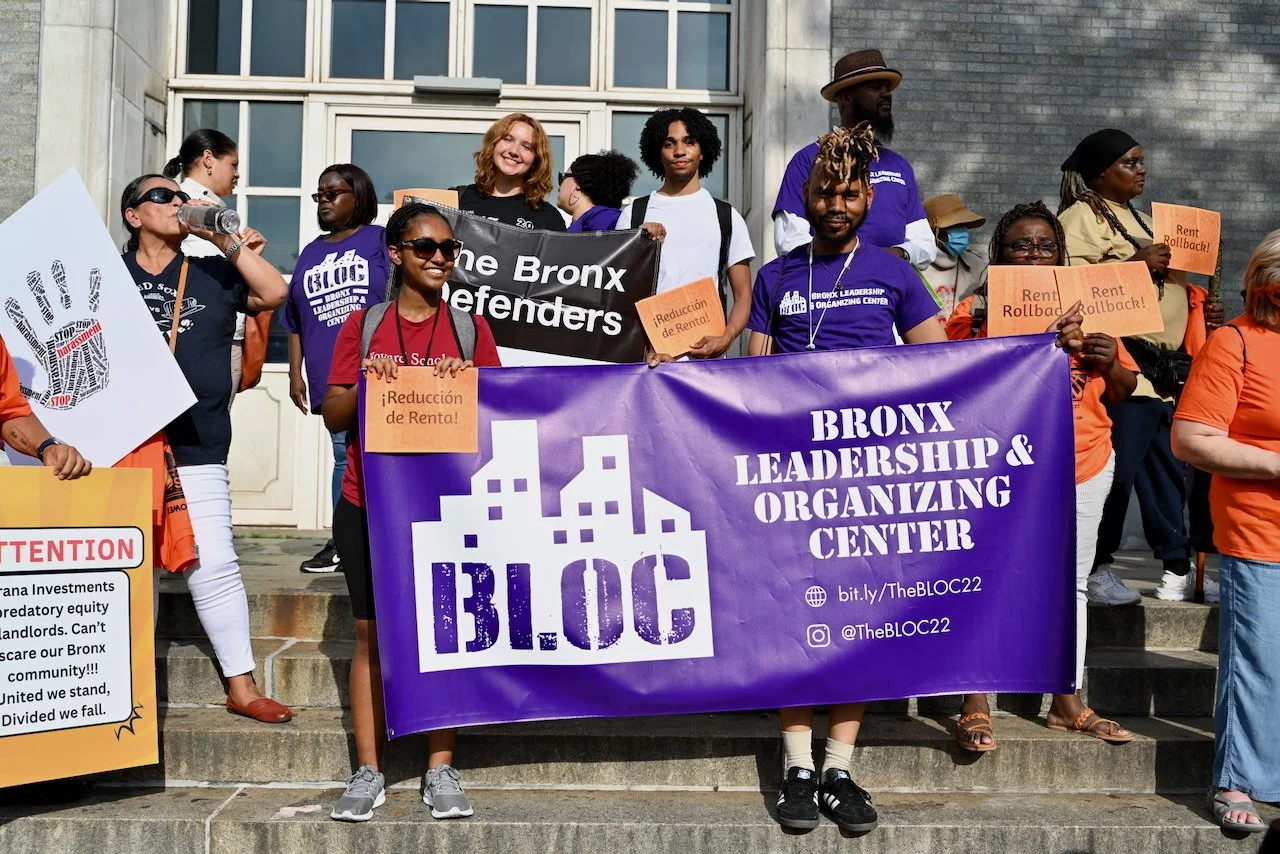 A diverse group of people gathered on stairs holding a purple banner that reads 'Bronx Leadership & Organizing Center' and a black sign saying 'the Bronx Defenders.' They are participating in a rally against rent increases, with some holding signs in Spanish and orange signs with messages about rent rollback and rent reduction. The background shows a building with large windows.