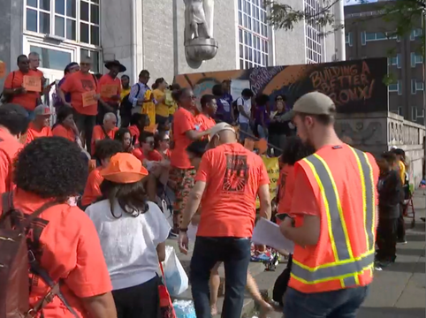 A group of people gathered outdoors for a rally, many wearing orange shirts and one person wearing a yellow safety vest, some holding papers, in front of a building with a banner that reads "Building a better Bronx."