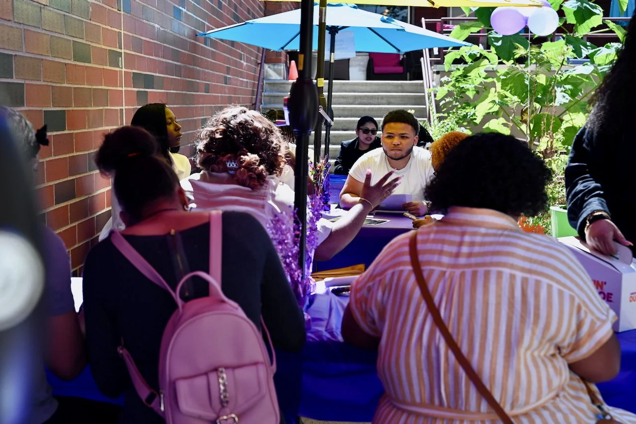 A diverse group of people sitting at a table outdoors under a blue umbrella, engaging in a discussion in an outside garden area.