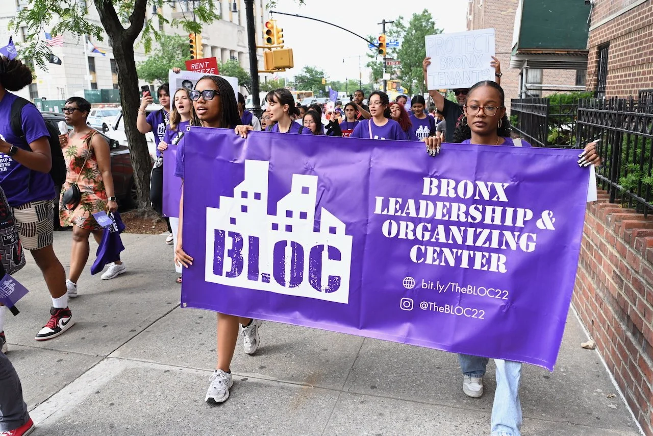 A group of people participating in a march. They are walking on a city sidewalk, holding a large purple banner that reads 'The Bronx Leadership & Organizing Center' and 'B.L.O.C.' with the organization's website and social media handles. Some signs are visible, including one that says 'Protect Bronx Tenants'. The participants are diverse, wearing purple shirts, and the scene is in an urban area with brick buildings and traffic lights.