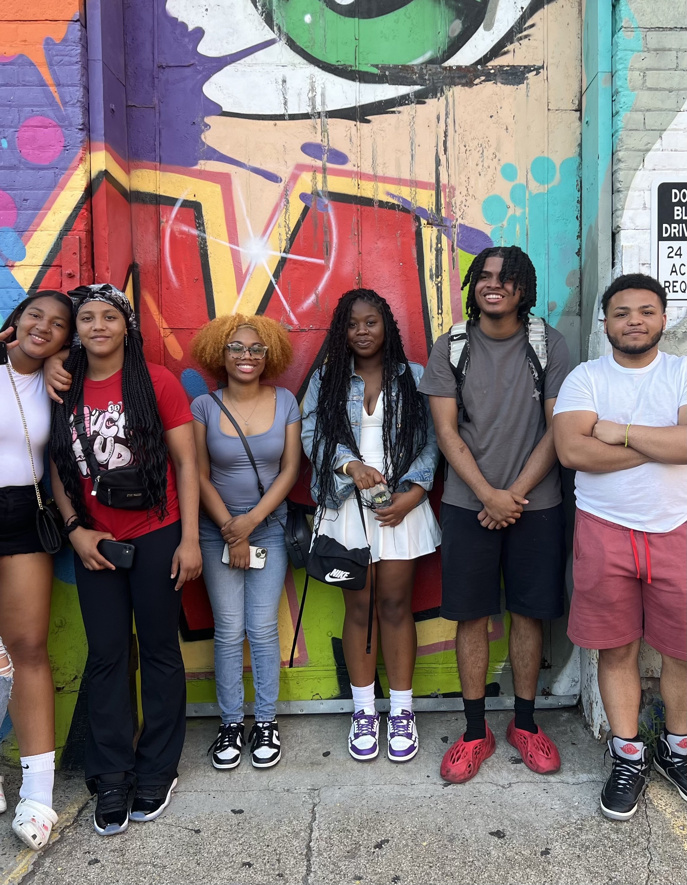 Group of six young adults standing in front of a colorful graffiti wall, smiling at the camera.