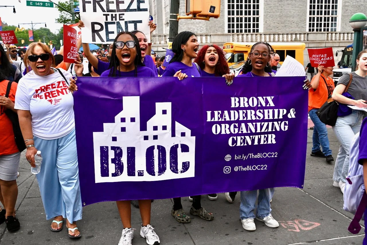 A group of people participating in a protest march, holding a large purple banner that reads "Bronx Leadership & Organizing Center BLOC" and "bit.ly/TheBLOC22," with other protesters holding signs that say "Freeze the Rent" and "Rent Freeze Now!"