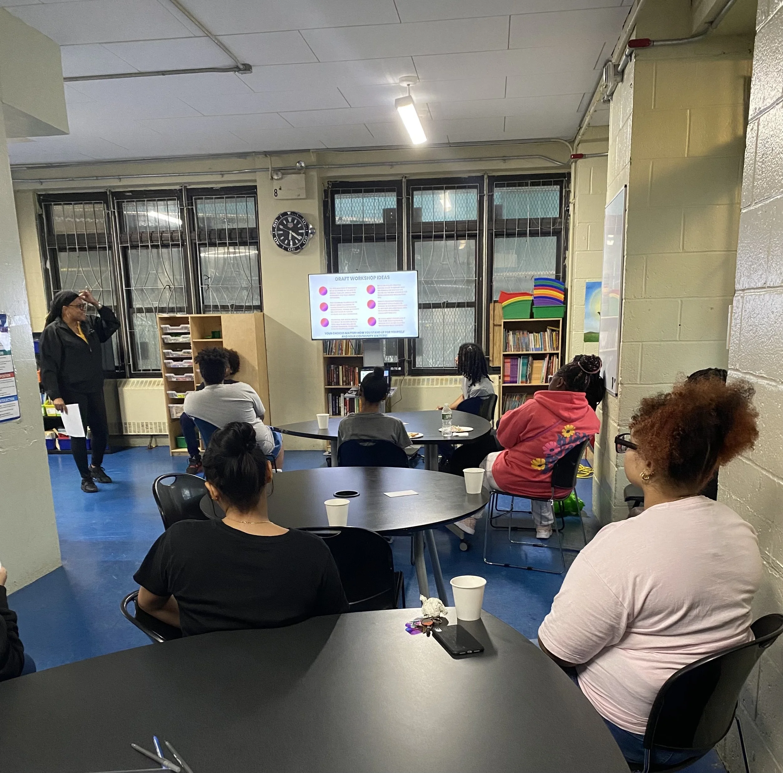 A group of people attending a workshop in a room with a facilitator presenting at the front. The room has black tables, some with cups, and books on shelves. A screen displays workshop ideas, and windows with bars are behind the presenter.
