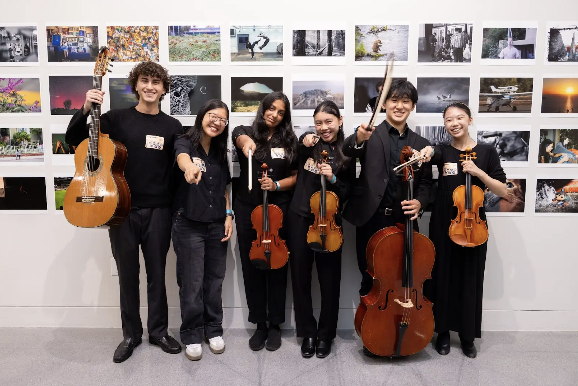 Group of seven young musicians holding string and wind instruments, standing in front of a wall of colorful photographs, all smiling and posing together.