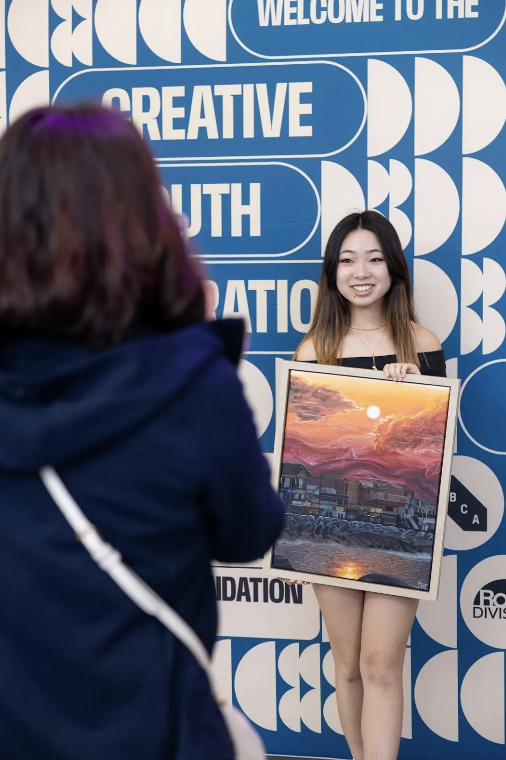 A young woman stands in front of a blue wall with white text and geometric designs, holding a colorful painting of a sunset over a body of water with mountains and buildings in the background. The text on the wall includes phrases like 'Welcome to th