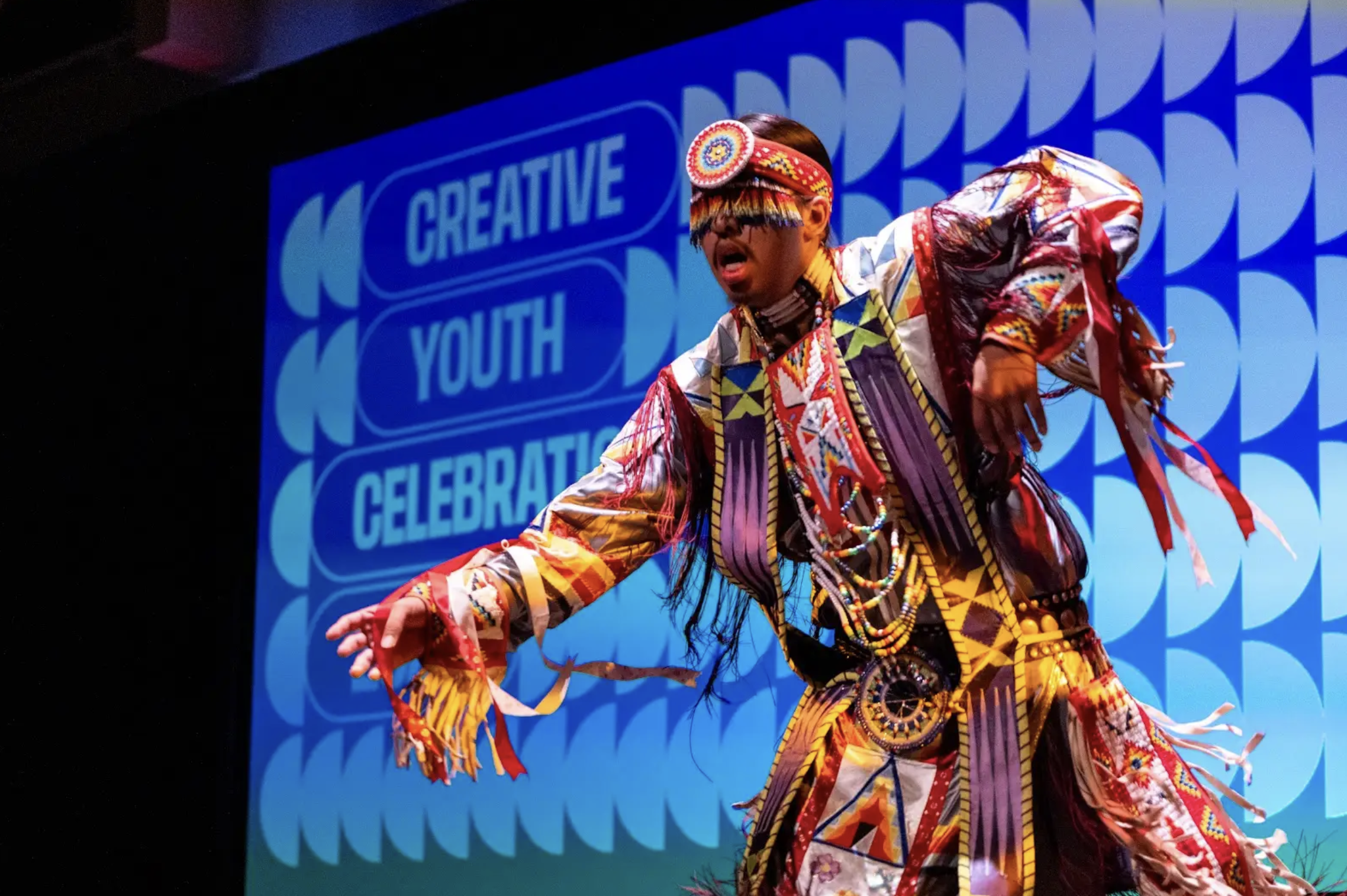 A performer dressed in colorful traditional tribal attire on stage during a cultural event, with a screen in the background reading 'Creative Youth Celebration'.