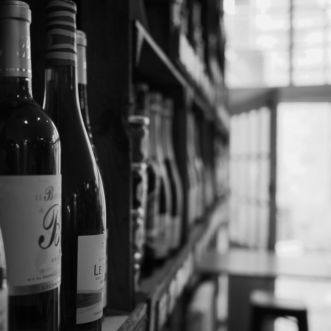 Black and white photo of wine bottles on a shelf in a store or cafe, with blurred background showing more bottles and furniture near a window.