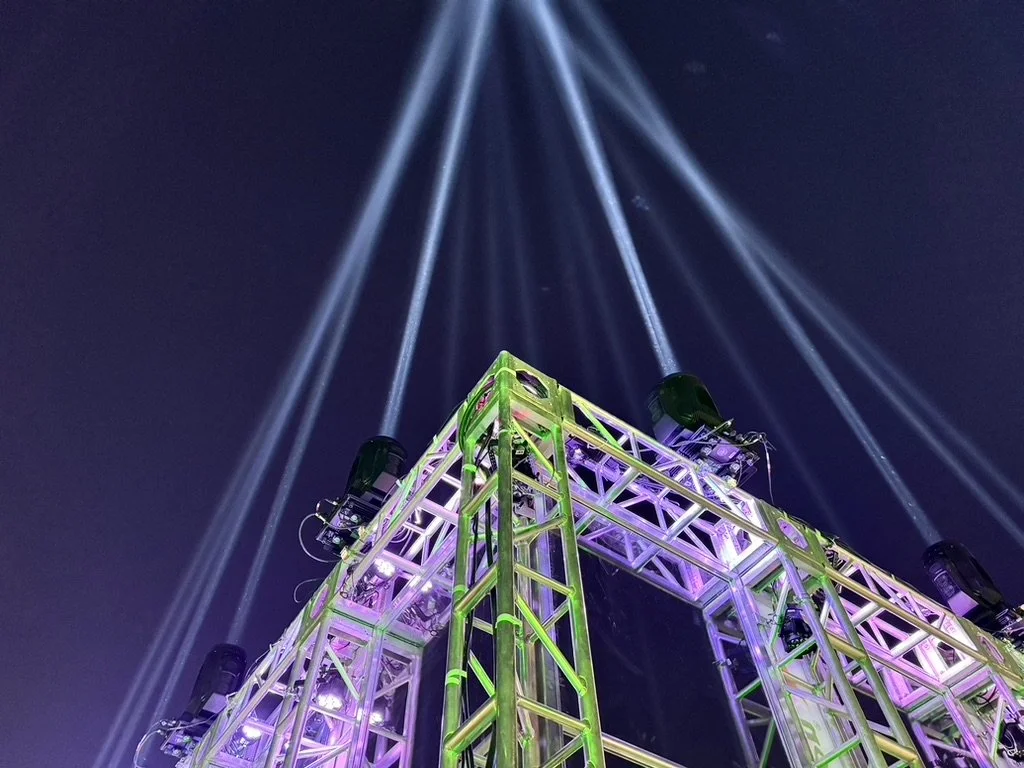 Light show with beams projecting into the night sky from a metallic stage structure illuminated with purple and green lights.