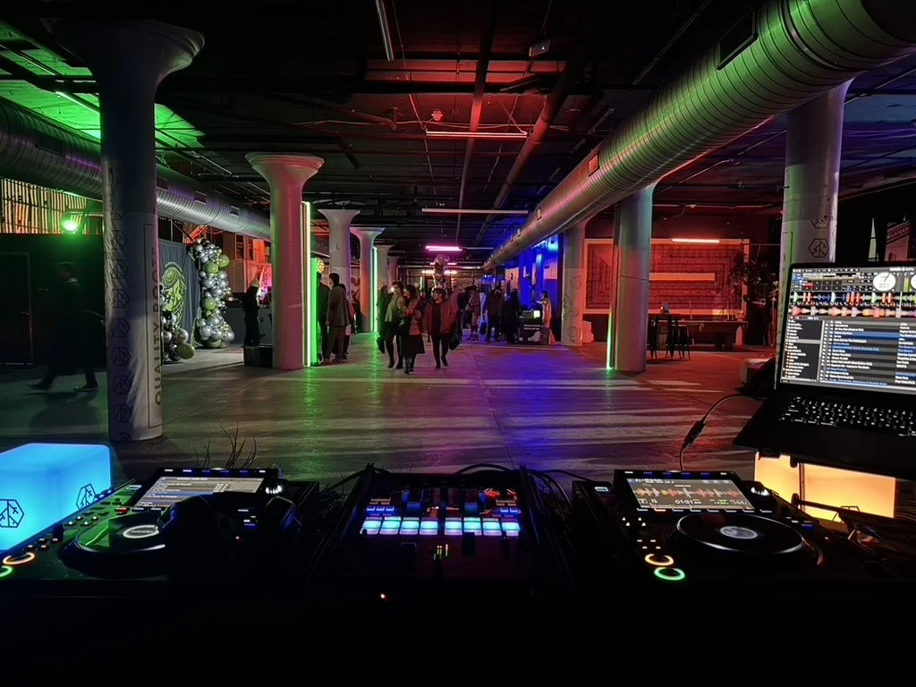Indoor nightclub with colorful lighting, people dancing, and a DJ setup with turntables and laptop.