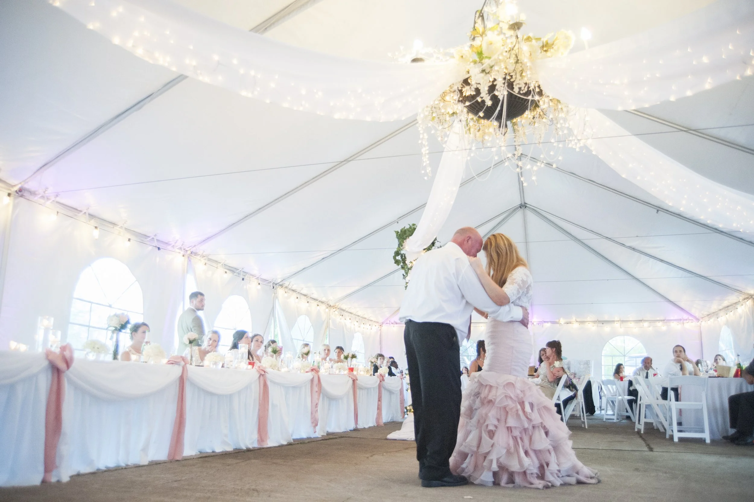 A bride and groom dancing at their wedding reception in a decorated tent, with guests seated at tables in the background.