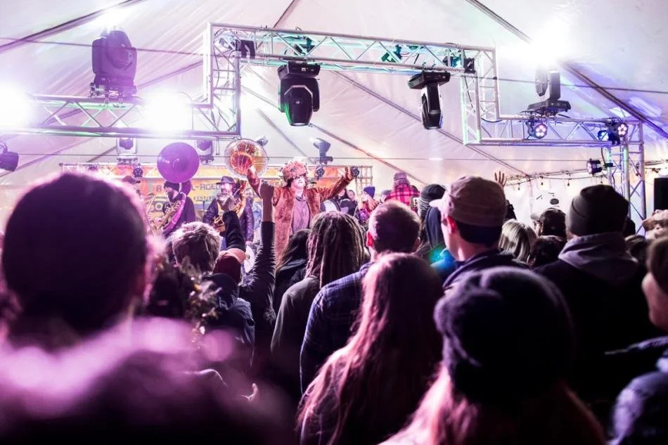 A lively concert scene inside a large tent with stage lighting, a performer wearing a hat singing or speaking to the crowd, and an audience engaging with raised hands.