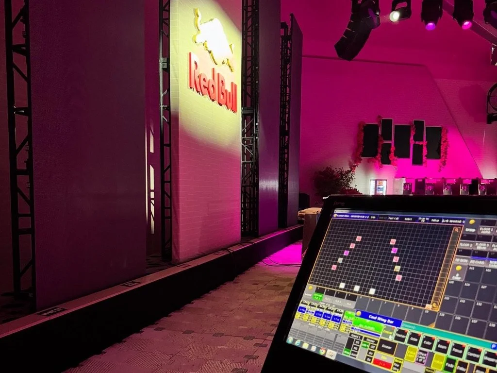 A concert stage with a Red Bull logo illuminated on a wall, pink and purple stage lighting, and a DJ or sound mixing console in the foreground.