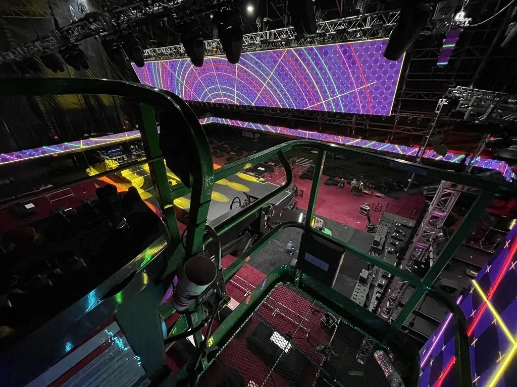Inside a large event venue with a stage featuring colorful, geometric LED patterns on a big screen and a view from a tall green lift, showing an empty floor with some equipment and few people.