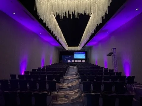 Conference room with rows of chairs facing a stage with a large screen, purple lighting, and elaborate chandelier ceiling design