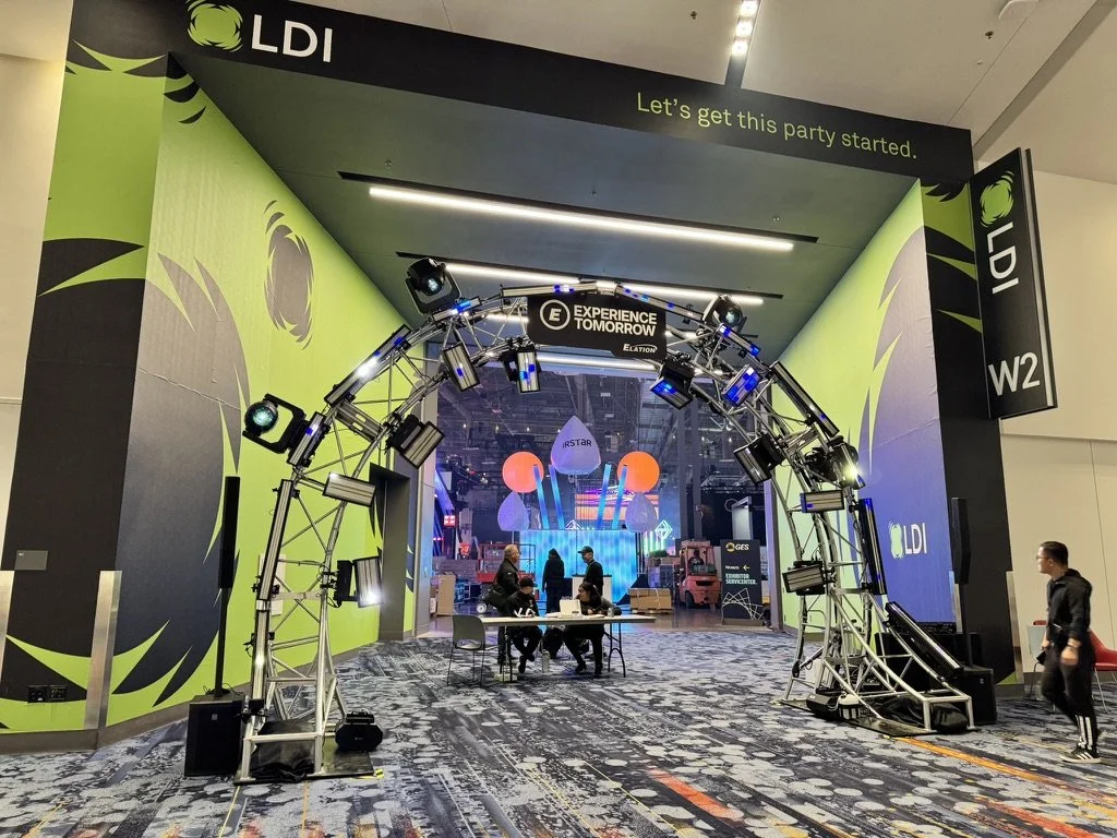 An event entrance with a large black and green archway inscribed with 'LDI' and 'Let's get this party started.' Inside, there is a stage with colorful lighting, abstract graphics, and people sitting at a table preparing for the event.