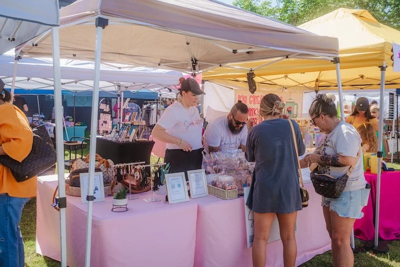 People shopping at an outdoor craft fair stall with pink tablecloths and yellow canopy, displaying various items and handmade crafts.